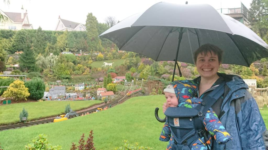 A person holding an umbrella stands outdoors, smiling while carrying a baby in a front-facing carrier. They are surrounded by a miniature landscape with small buildings and greenery in the background.